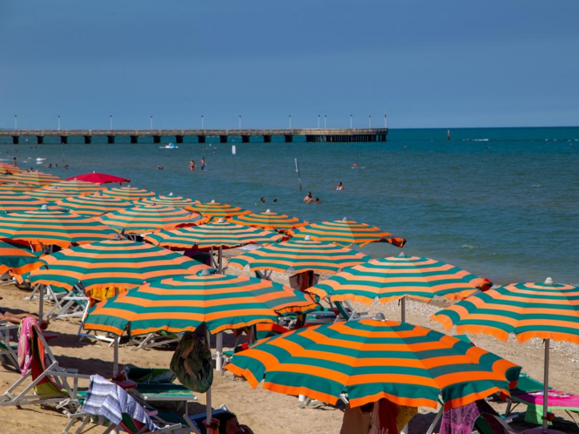 Spiaggia vivace con ombrelloni arancioni e verdi, mare calmo e pontile in lontananza.