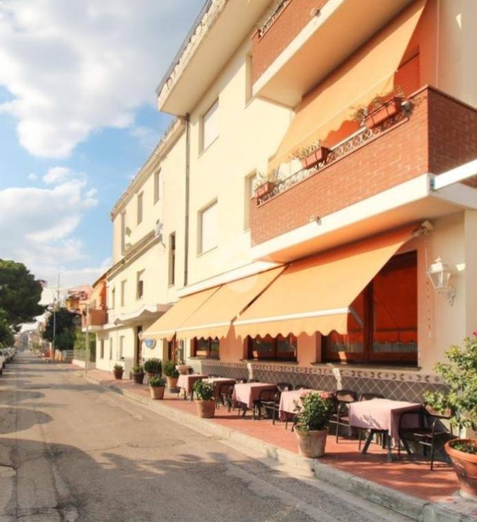 Strada tranquilla, tavolini all'aperto, tende arancioni, atmosfera accogliente e soleggiata.