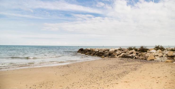 Spiaggia serena, onde leggere, cielo azzurro e nuvole soffici sopra un molo roccioso.