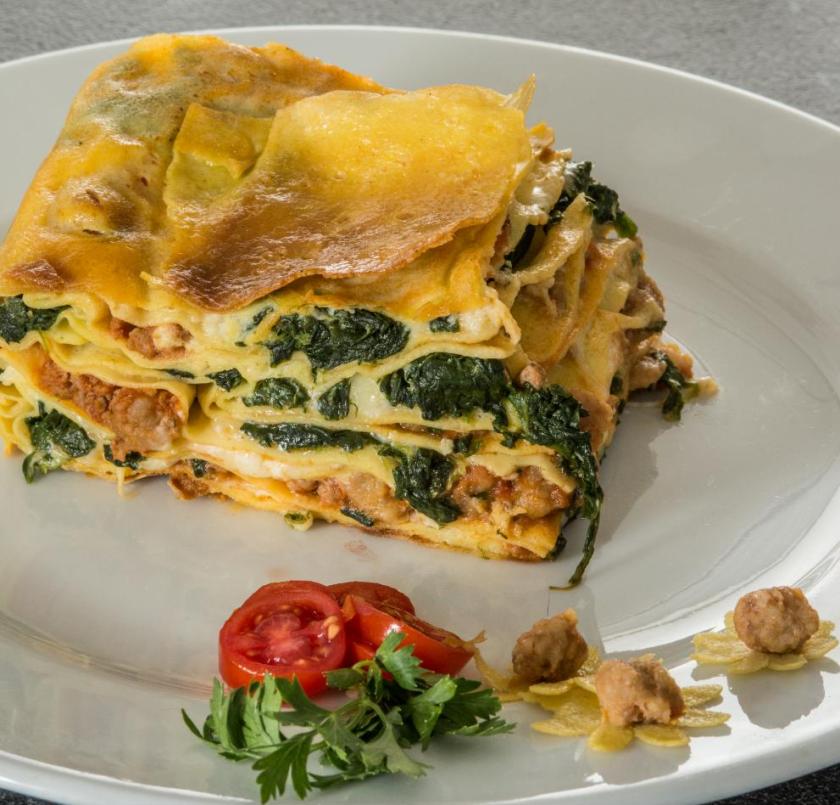 Lasagna dorata e succulenta con spinaci, carne, pomodorini e prezzemolo su piatto elegante.